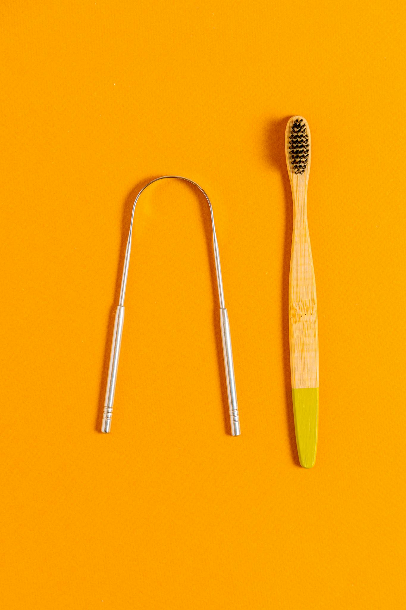 A bamboo toothbrush and tongue scraper on a vivid orange surface for sustainable oral hygiene.