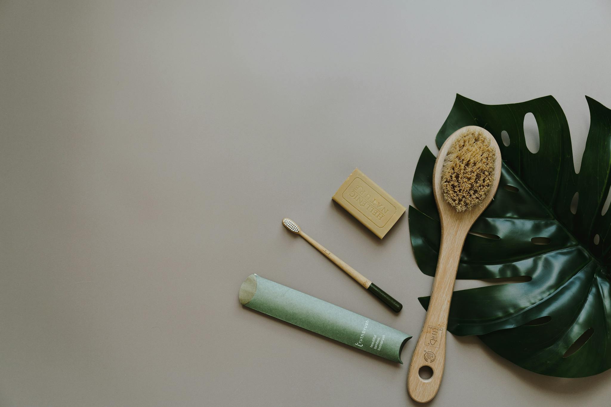 Flat lay of sustainable bath products including soap, brush, and eco toothbrush on a leaf background.