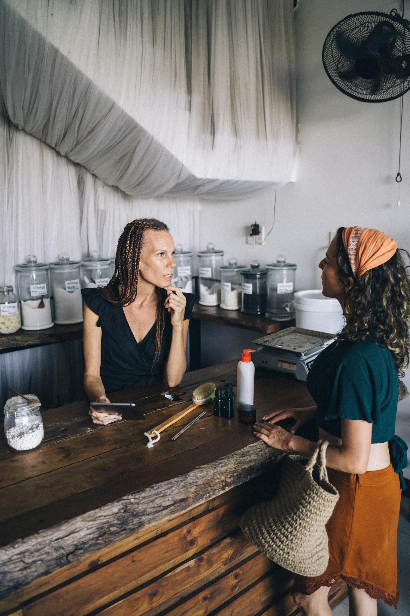 Two women engaging in conversation at an eco-friendly refillable shop counter.